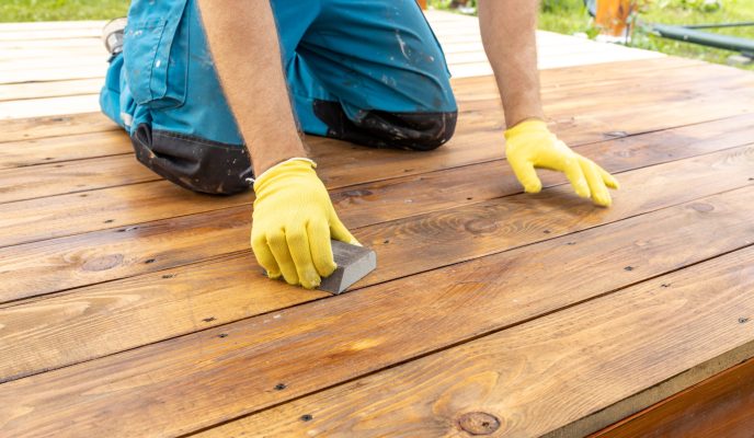 a close up view of a person wearing yellow gloves sanding a wooden deck with a block sander. the deck is made of weathered wood planks, and the person is kneeling on the deck.